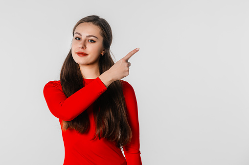 Confident young woman points finger right, showing news, promo banner, offer. Beautiful girl in casual red clothes looks serious, stands against neutral studio background