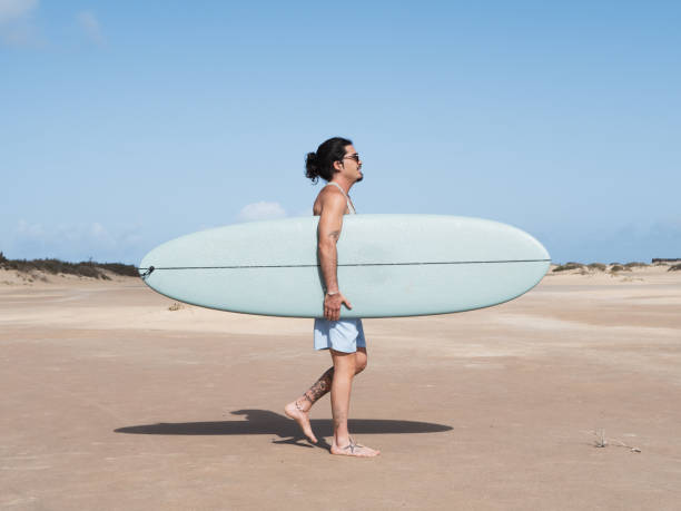 A man stands on a sandy beach, holding his surfboard, gazing towards the ocean waves. The atmosphere evokes feelings of excitement and connection with nature, representing a passion for the surfing lifestyle.
