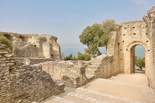 Wide-angle view of the residential area in the archaeological complex of Grotte di Catullo, a Roman villa supposedly built by Latin poet Gaius Valerius Catullus between the end of the 1st century BC and the beginning of the 1st century AD at the northernmost end of the Sirmione peninsula, on the shore of Lake Garda. A perfectly clear sky, the warm, oblique light of a late afternoon, picturesque olive trees. A stretch of Lake Garda and its western shore can be seen at the distance. Palette based on creamy yellow tones and pale cyans, with green accents. High level of detail, natural rendition, realistic feel. Developed from RAW.