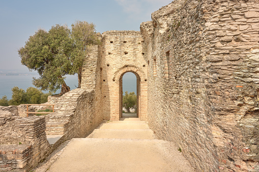 Wide-angle view of a long flight of steps and a tall portal in the archaeological complex of Grotte di Catullo, a Roman villa supposedly built by Latin poet Gaius Valerius Catullus between the end of the 1st century BC and the beginning of the 1st century AD at the northernmost end of the Sirmione peninsula, on the shore of Lake Garda. A perfectly clear sky, the warm, oblique light of a late afternoon, picturesque olive trees. A stretch of Lake Garda and its western shore can be seen at the distance. Palette based on creamy yellow tones and pale cyans, with green accents. High level of detail, natural rendition, realistic feel. Developed from RAW.