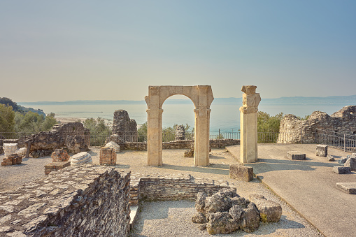 Wide-angle view of the Arco Imperiale (Imperial Arch) in the archaeological complex of Grotte di Catullo, a Roman villa supposedly built by Latin poet Gaius Valerius Catullus between the end of the 1st century BC and the beginning of the 1st century AD at the northernmost end of the Sirmione peninsula, on the shore of Lake Garda. A perfectly clear sky, the warm, oblique light of a late afternoon, picturesque olive trees, the remains of columns and pillars lying on the ground. A long stretch of Lake Garda and its western shore can be seen at the distance. Palette based on a wide range of rich earthy tones, creamy yellows and pale cyans, with green and orange accents. High level of detail, natural rendition, realistic feel. Developed from RAW.