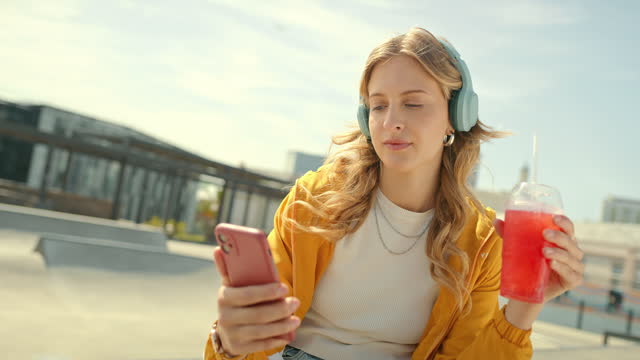 Young woman with headphones and juice, listening to music in a urban city skatepark for gen z lifestyle. Face of a student drinking red slushy with smartphone and audio streaming on 5g technology
