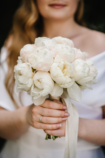 A young girl is standing and holding a bouquet of white peonies with a ribbon at a wedding ceremony. Flowers close up. Engagement ring.
