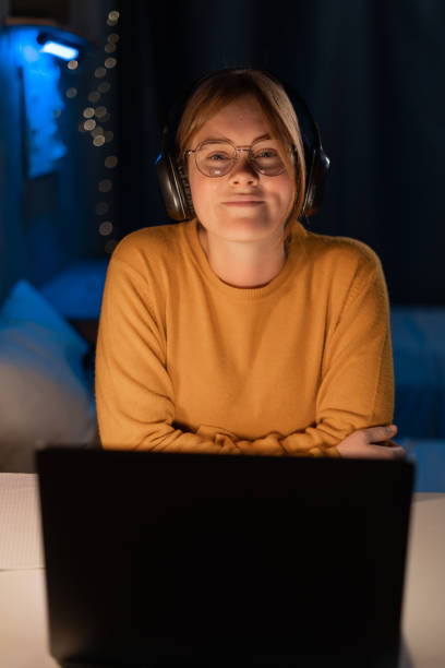 istock Young female student sitting at the table, using headphones when studying in dormitory at late evening looking at camera, close up. 1472280402