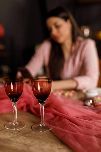 Selective focus shot of young woman sitting at table that she set up and decorated for Valentine's day celebration, using smart phone while waiting for her loved one. Focus on foreground.