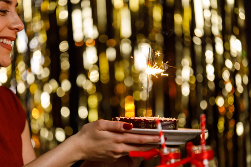 Side view of happy young woman sitting against gold colored tinsel curtain, holding a heart shaped cake with sparklers inside burning and making a wish while celebrating her birthday.