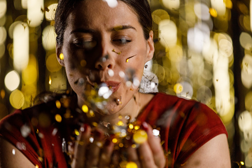 Candid shot of happy young woman standing against gold colored tinsel curtain, having fun while blowing confetti towards camera during a celebratory party.