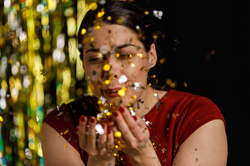 Candid shot of happy young woman standing against gold colored tinsel curtain, having fun while blowing confetti towards camera during a celebratory party.