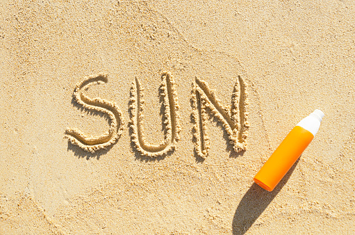 word Sun is written on the sea sand. Orange bottle of sunscreen and the word Sun on the beach sand. Template, layout, text space. Summer background with UV protection lotion on the sea coast