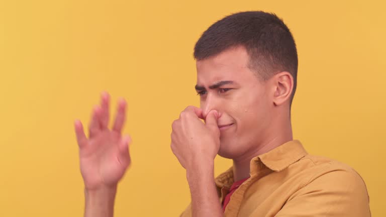 Smelly, stink and bad with a man waving his hand in studio on a yellow background to remove a foul odor. Nose, disgust and smelling with a young male on a pastel wall background trying not to breath