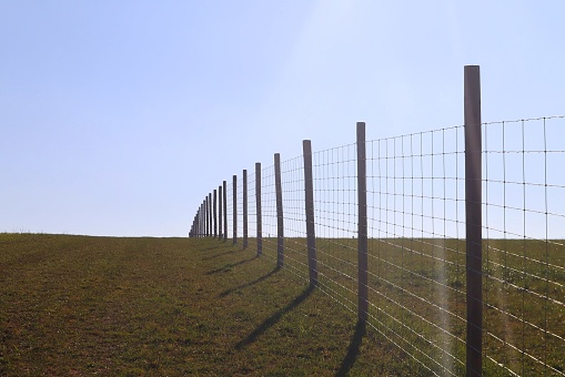 Wire fence in a field marking boundary on farmland with blue sky on a hill, farmland all around with the fence being made of wooden posts and copy space in the empty sky