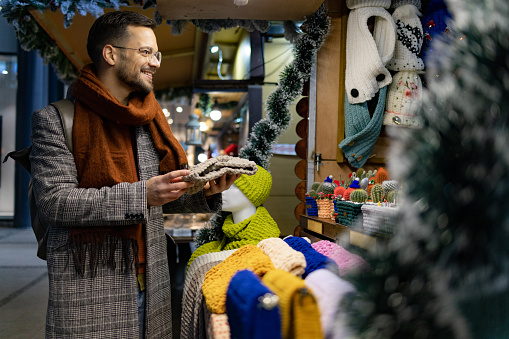 Happy male tourist choosing knitted headwear at market stall.