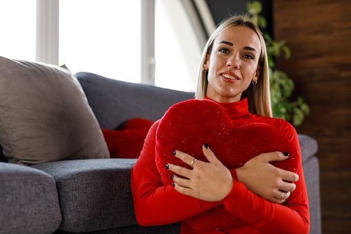 Portrait of happy young woman in love sitting on the living room floor, by the sofa, embracing a heart shaped pillow and daydreaming of her loved one. She is looking at camera and smiling joyfully.