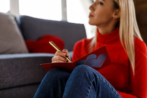 Portrait of happy young woman sitting on the living room floor, by the sofa, contemplating while writing a Valentine card for a loved one.