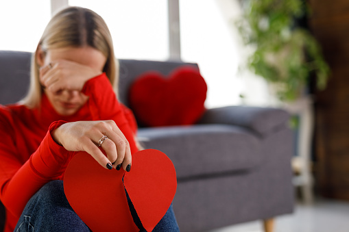 Selective focus shot of sad young woman sitting on the floor, covering her eyes while crying and holding a broken heart made out of paper.