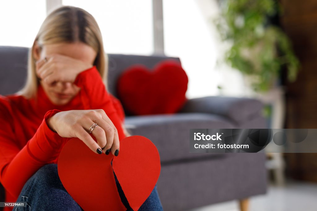 Sad young woman holding a broken heart made out of paper and crying Selective focus shot of sad young woman sitting on the floor, covering her eyes while crying and holding a broken heart made out of paper. Heart Shape Stock Photo