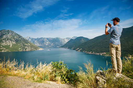 Tourist man taking a picture on his smartphone, standing on the edge of a cliff, enjoying amazing sea view of the Bay of Kotor, Montenegro