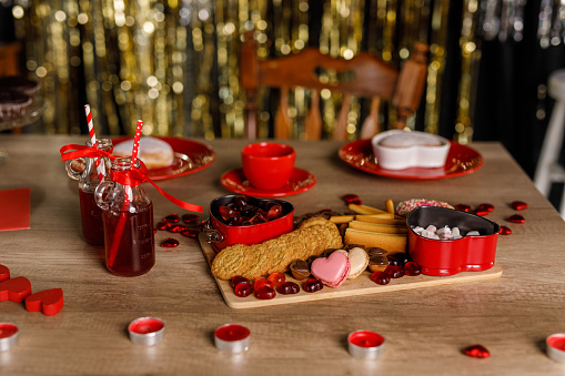 Close up shot of a delicious sweet charcuterie board filled with cookies, heart shaped candy and macaroons, set up on the table next to red colored drinks for a Valentine's day celebration.