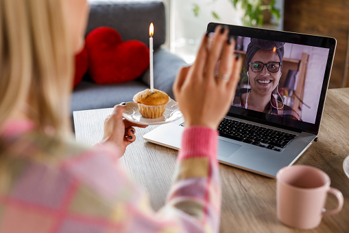 Over the shoulder view of unrecognizable young woman making a wish before blowing out the birthday candle in a chocolate chip muffin while video conferencing a friend with whom she is virtually celebrating her birthday.