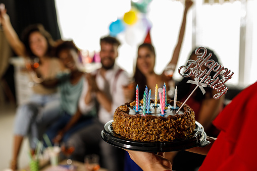 Selective focus shot of unrecognizable young woman bringing out the cake with birthday candles during a friend's birthday party celebration. Group of diverse friends hanging out and having fun in the background.