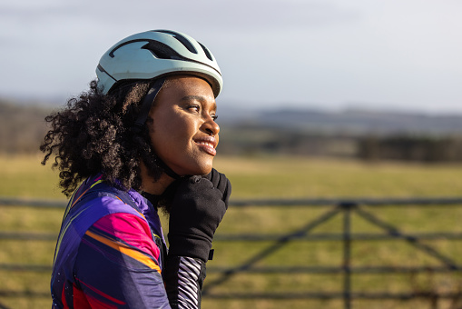 Woman riding her bike in a rural area in the countryside, staying active in the North East of England. She is taking a break from cycling, standing near a gate to a field enjoying the scenery. She is taking off her cycling helmet.