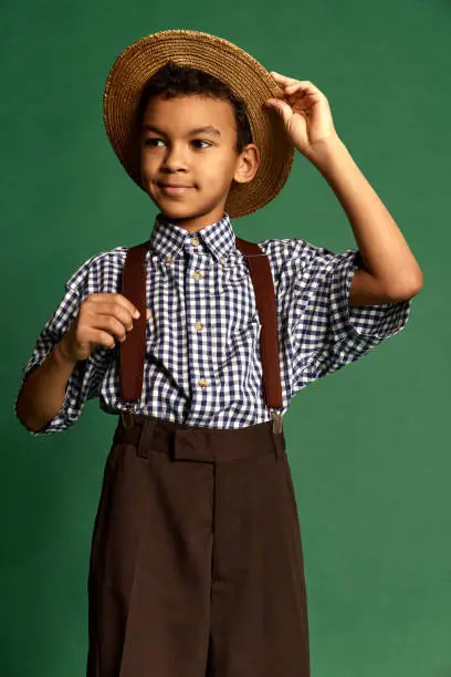 Kid in straw hat. Cute handsome little boy, child in checkered shirt and suspender shorts isolated over dark green background. Concept of childhood, friendship, retro fashion style