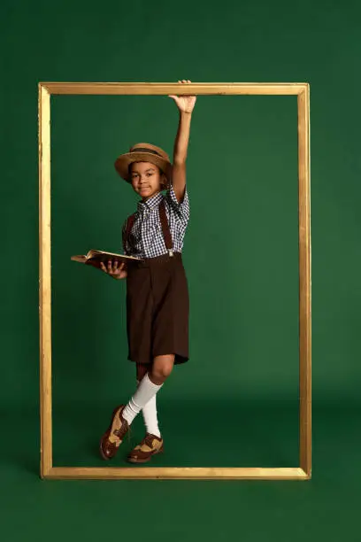 African little boy, child in checkered shirt and suspender shorts standing near huge picture frame over dark green background. Concept of childhood, art, friendship, retro fashion style