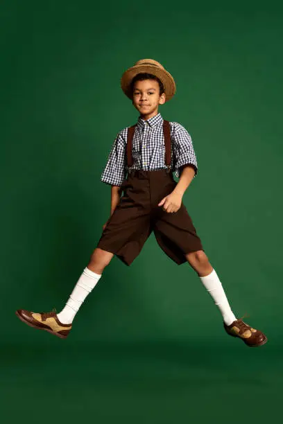 Happy little boy, child in checkered shirt and suspender shorts jumping, having fun over dark green background. Concept of happiness, childhood, friendship, retro fashion style