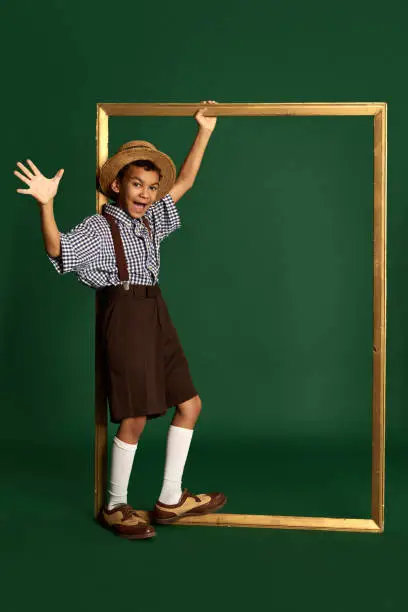 African little boy, child in checkered shirt and suspender shorts standing near huge picture frame over dark green background. Concept of childhood, art, friendship, retro fashion style