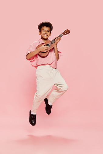 Happy emotional little african boy in retro style clothes and cap posing with ukulele guitar isolated on pink background. Concept of music, childhood, education, fashion and aspiration
