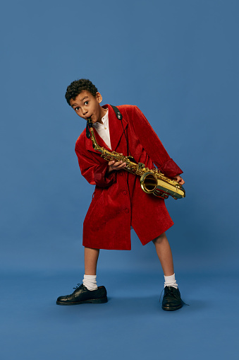 Like jazz man. Portrait of cute little african boy wearing huge man's jacket and shoes playing on saxophone over blue background. Fashion, art, music and creative style. Looks happy, cheerful