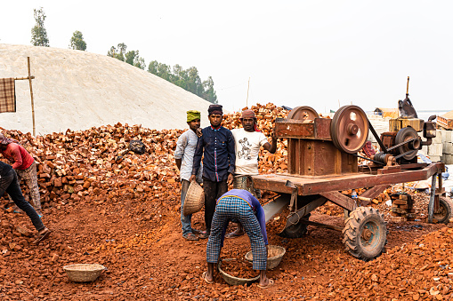 Gaibandha, Babgladesh-January 23th 2022-Three men looking at a camera lens. Six workers prepared for work with a crusher machine. Bricks are crushed by a crusher machine and men carry broken bricks on top of their heads.
