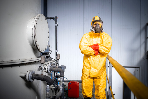 Portrait of factory worker in protective yellow suit, gas mask and gloves holding arms crossed inside chemicals production plant.