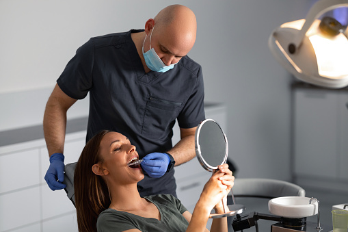 Shot of a young woman looking at the hand mirror, checking her results in the dentist's office while the dentist is removing her transparent trainer dental braces.
