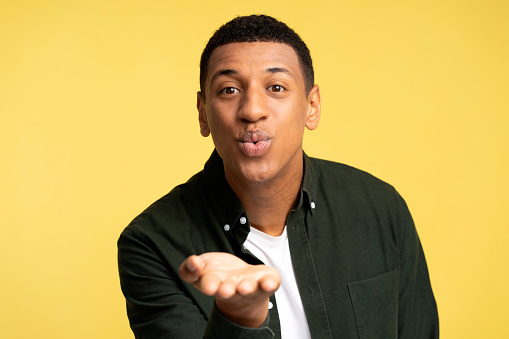 Portrait of amorous handsome brunette man holding palms up while sending air kiss at camera, demonstrating feelings. Indoor studio shot isolated on yellow background