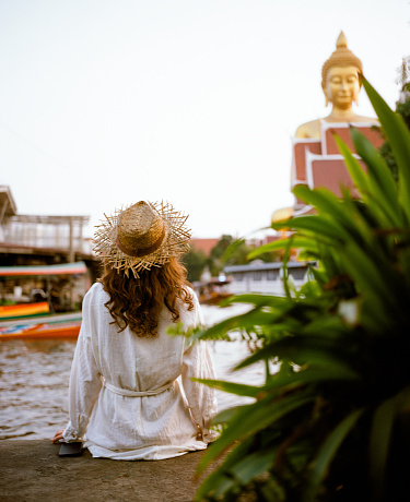 Young Caucasian woman in straw hat and white dress sitting on the riverbank in Bangkok and looking at huge statue of Golden Buddha