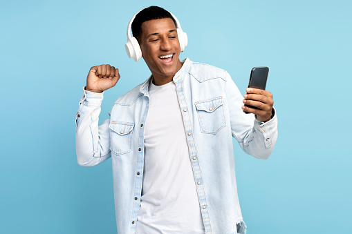 Multiracial man wearing headphones using smartphone, listening music and dancing. Happy guy with earphones holds phone, isolated on blue