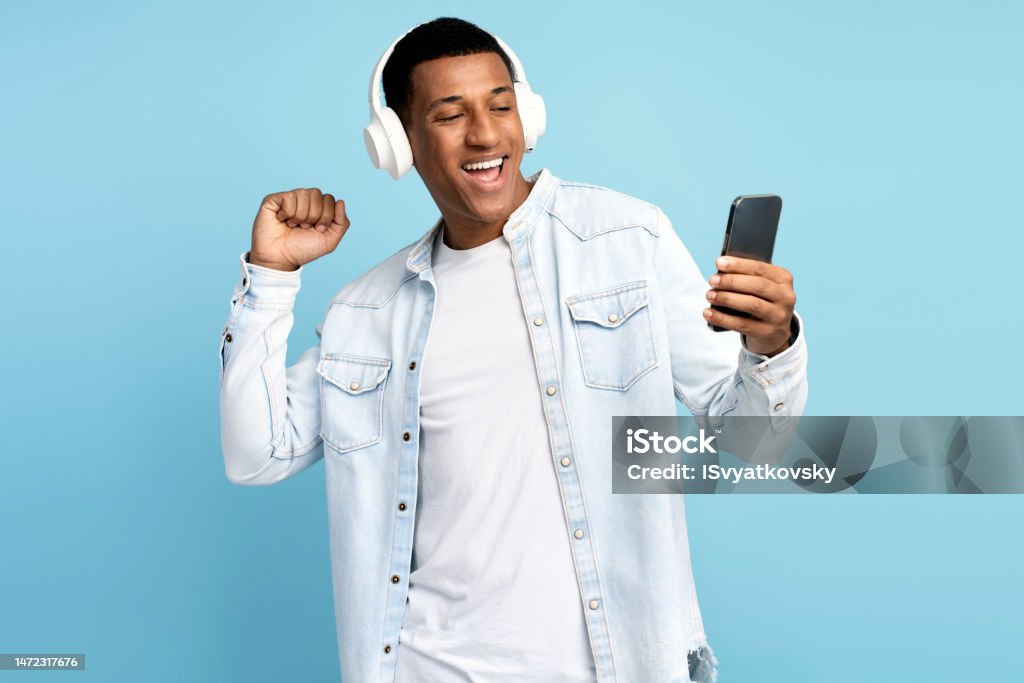 Multiracial man wearing headphones using smartphone, listening music and dancing Multiracial man wearing headphones using smartphone, listening music and dancing. Happy guy with earphones holds phone, isolated on blue Headphones Stock Photo