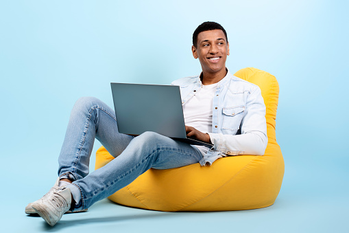 Happy man looking away, typing information content, using laptop for working, sitting at the armchair isolated over blue background. Search in the internet concept