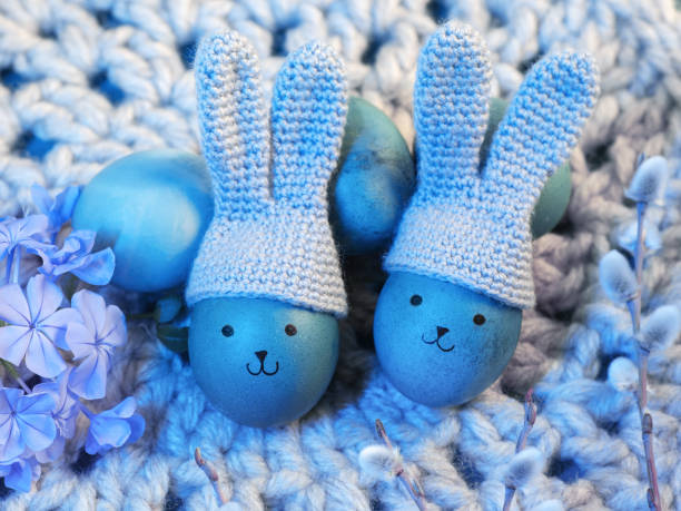 Easter eggs in knitted hats with bunny ears and painted faces. Knitted napkin, blue background