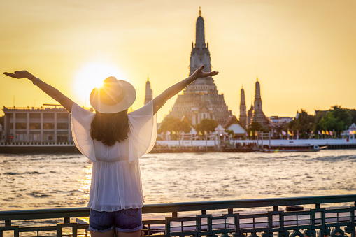 A happy tourist woman looks at the famous Wat Arun temple in Bangkok, Thailand, during golden sunset time