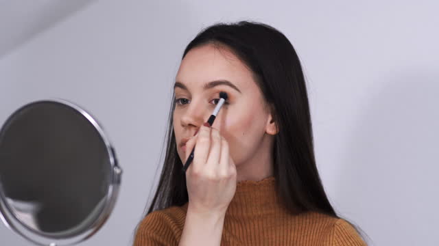 Portrait of a young woman applying eye shadow with brush looking in circular mirror