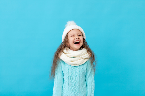 laughing little girl child in winter clothes hat and sweater on a blue isolated background smiling and having fun, a place or space for text
