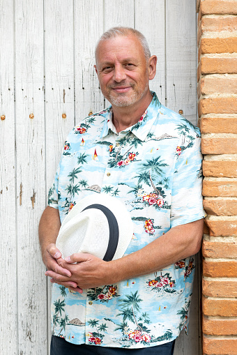 A waist-up portrait of a male mid-adult smiling and looking into the camera. He is on holiday standing in front of a wooden door holding his sun hat with both hands. He is located near Toulouse in France.
