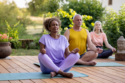 A full shot of a group of mature adults on a wellness retreat in France. They sit with their legs crossed and eyes closed outdoors meditating together. The retreat is located near Toulouse in France.
