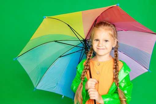 a child girl on an isolated green background with an umbrella and a raincoat smiles, space for text