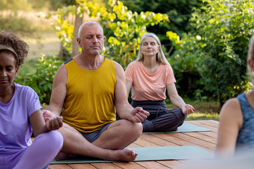 A group of mature adults on a wellness retreat in France. They sit with their legs crossed and eyes closed outdoors meditating together. The retreat is located near Toulouse in France.