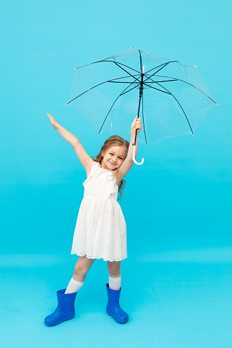 Happy cute little girl in blue rubber boots and a cotton white dress holding an umbrella on a blue background in the studio and smiling and fooling around, space for text