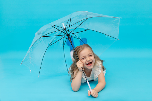 A happy cute little girl in blue rubber boots and a cotton white dress holding an umbrella lying on a blue background in the studio laughing, smiling and fooling around, a place for text