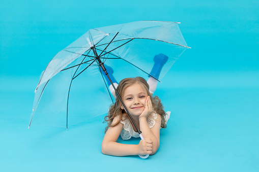 A happy cute little girl in blue rubber boots and a cotton white dress holding an umbrella lying on a blue background in the studio laughing, smiling and fooling around, a place for text